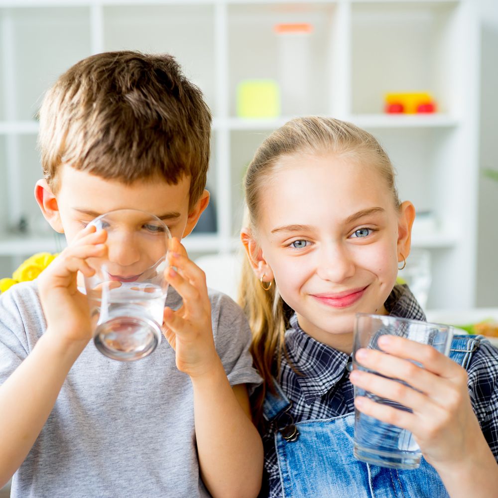 A young boy drinks purified water from a reverse osmosis system while a smiling girl holds a glass next to him.