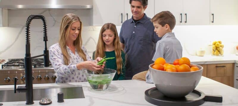 A family standing in a kitchen as a woman shows a young girl how to wash green beans in a bowl.