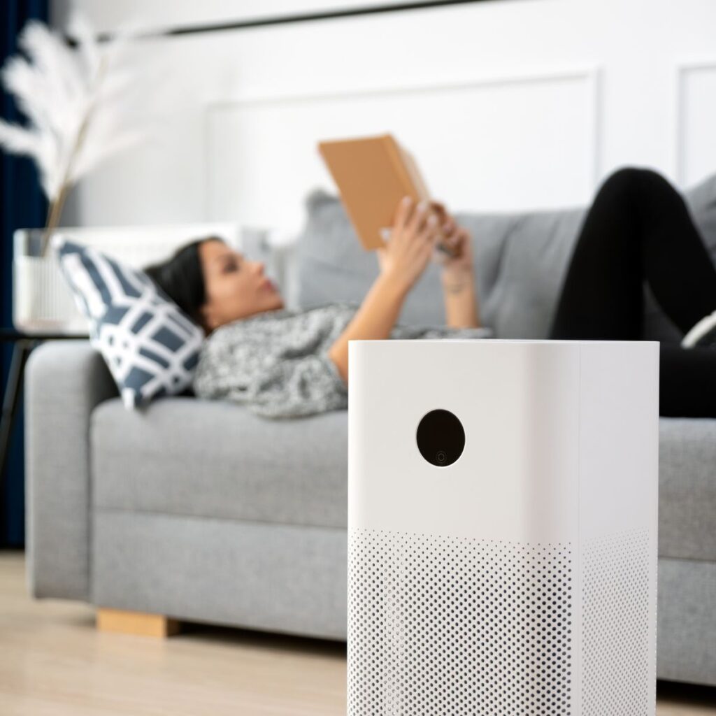young woman laying down on the couch in the background reading a book as a white air purifier sits in front of her