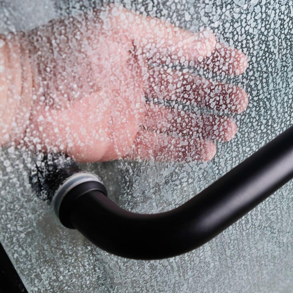 A hand rests against a shower door covered in water spots near a black handle, an image often seen in our frequently asked questions.