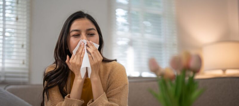 A woman sits on a sofa, blowing her nose into a tissue.