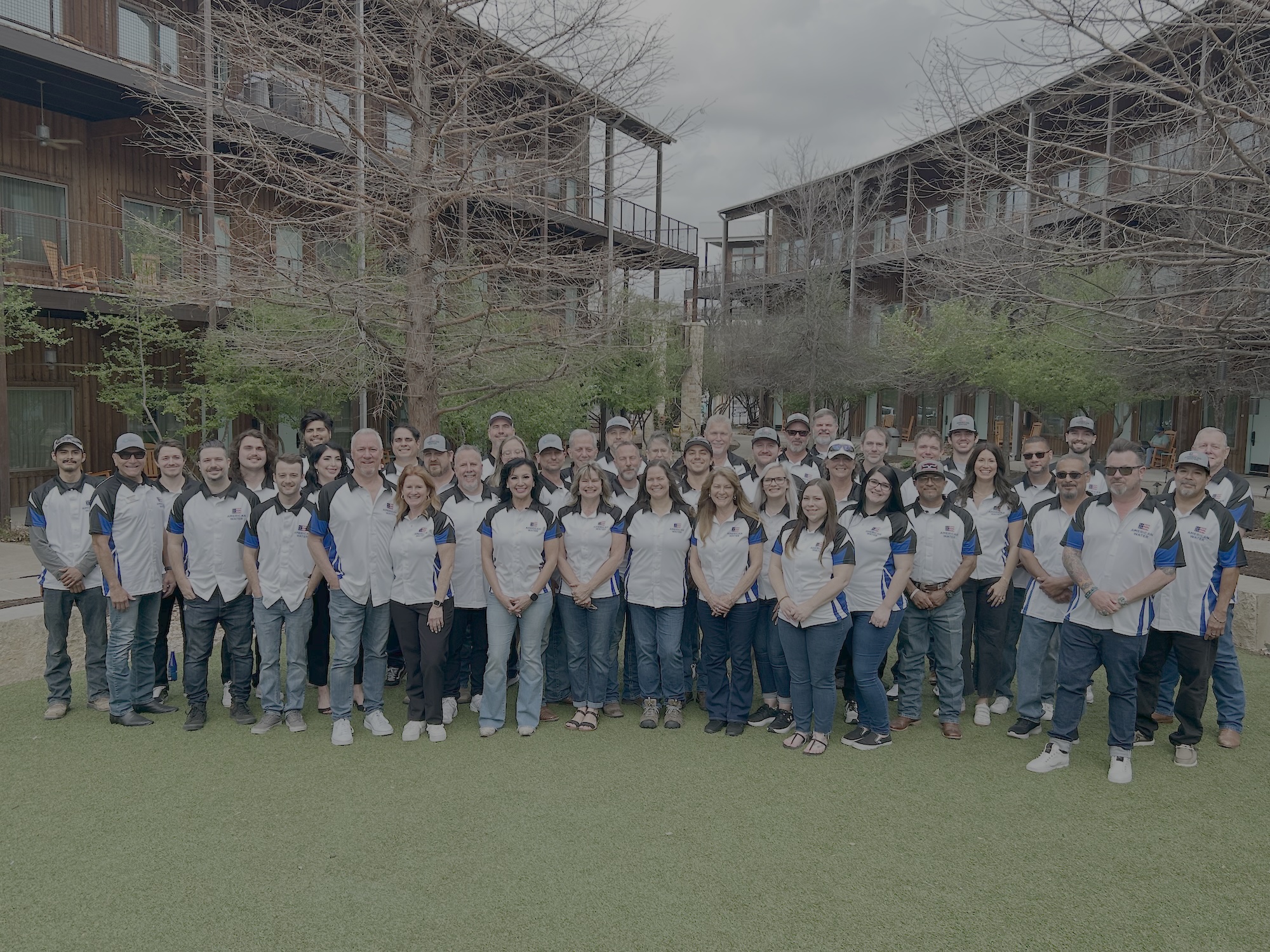 A large group of people in matching white polo shirts with blue accents stand together on a grassy lawn in Austin, in front of modern, multi-story buildings that utilize water filtration.