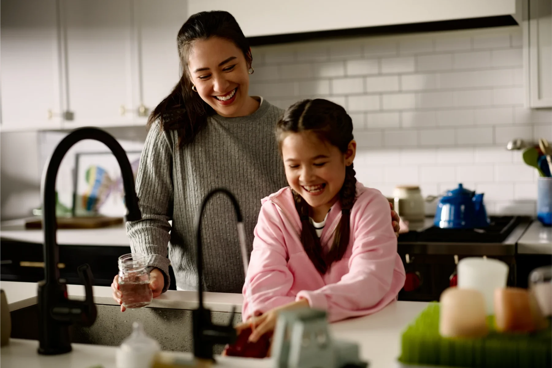 A smiling woman and girl laughing together while washing dishes at a kitchen sink.