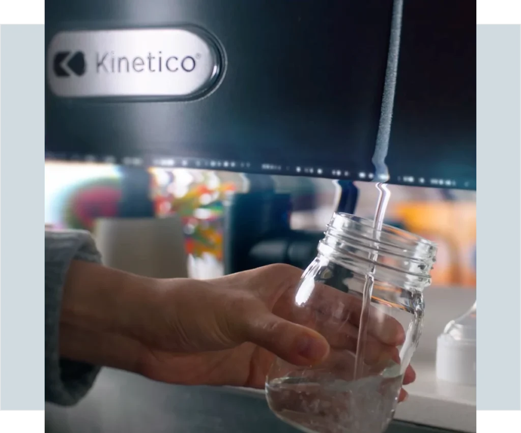 A hand holds a glass jar under the tap of a Kinetico water filtration system.